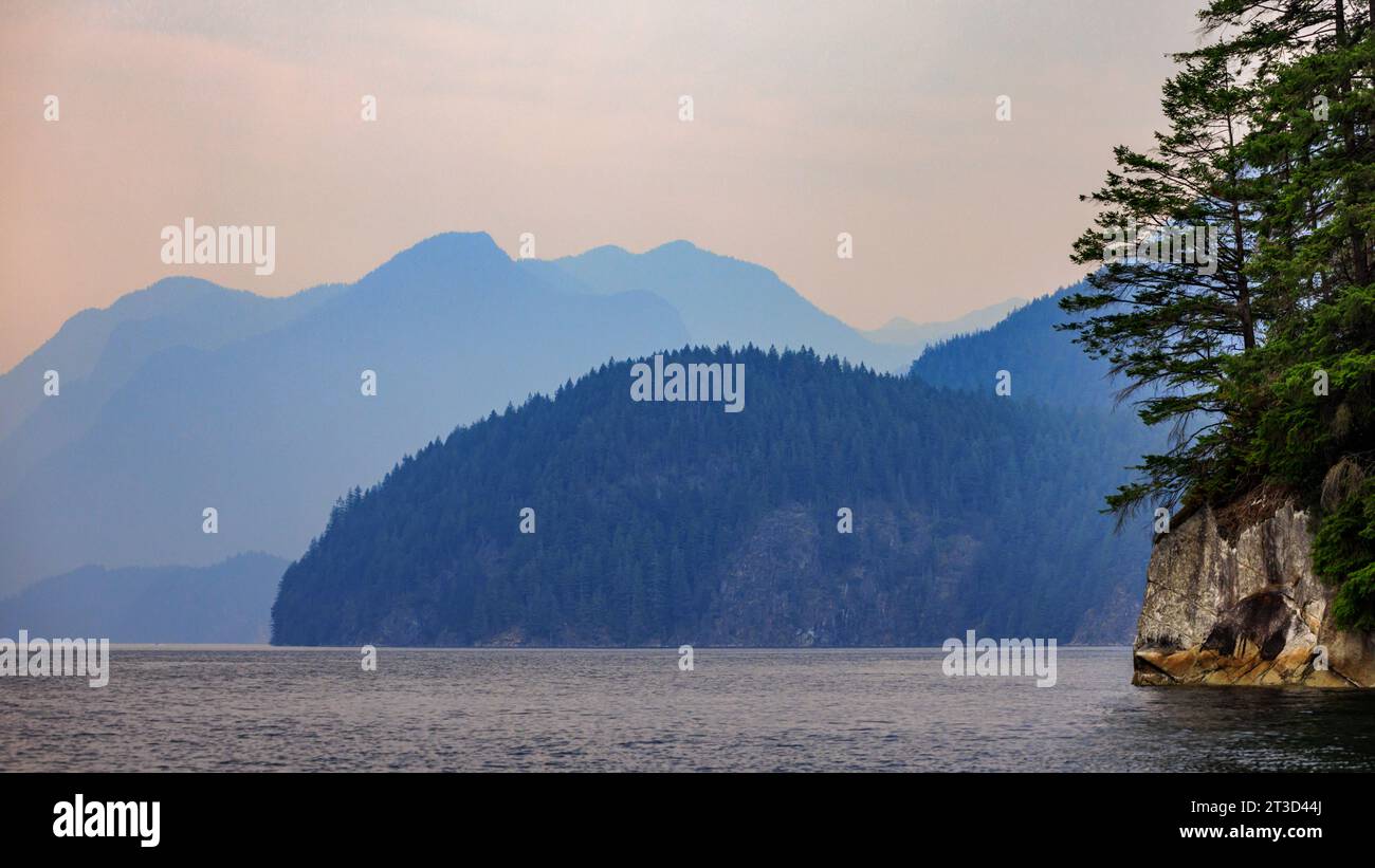Le montagne nel remoto braccio Indiano svaniscono nel cielo rese arancioni dal fumo di fuoco nella Columbia Britannica, Canada. Vista del paesaggio da una barca sul fiordo. Foto Stock