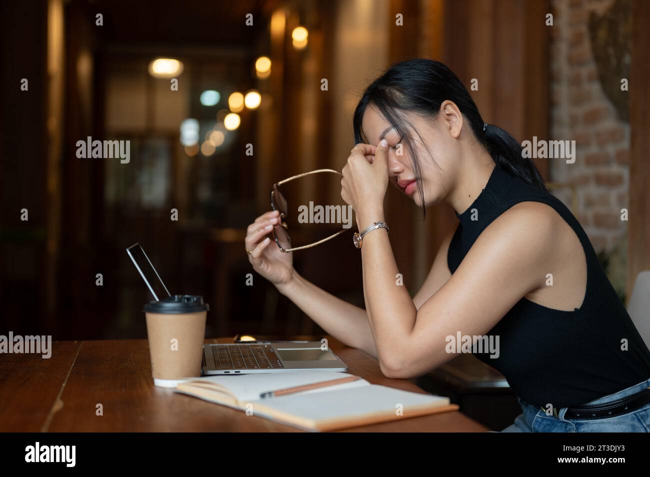 Una donna asiatica stanca e sovraccarica si sta togliendo gli occhiali da vista, sentendo affaticamento per gli occhi dopo aver guardato lo schermo per troppo tempo mentre lavorava sul suo compito Foto Stock