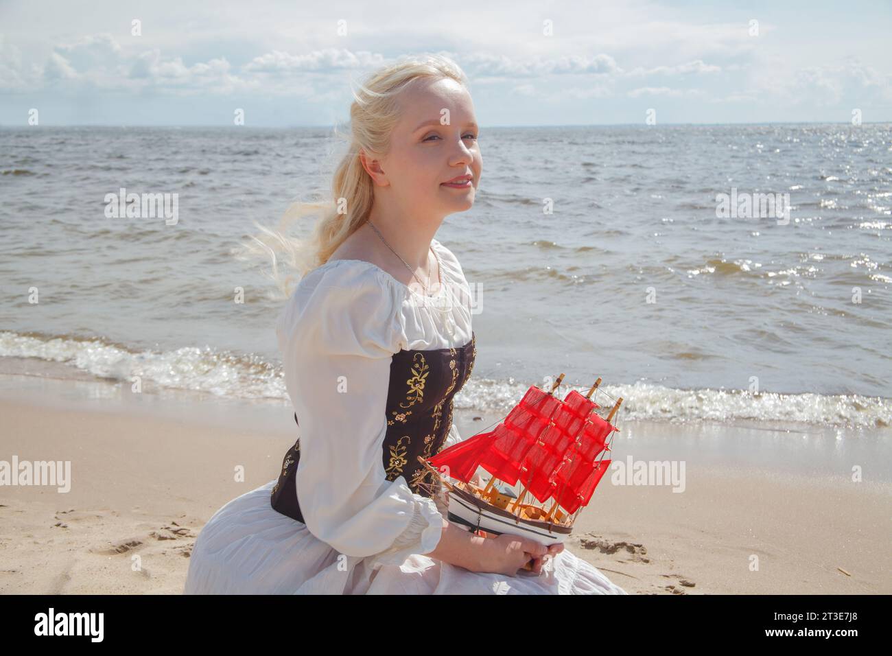 Ritratto di una giovane e romantica modella bionda in abito retrò che regge una barca a vela rossa sulla spiaggia di mare Foto Stock
