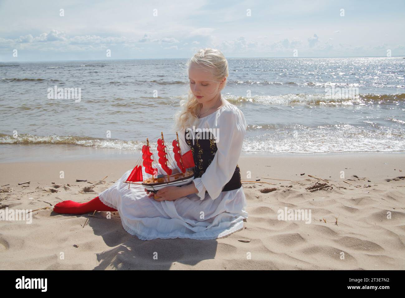 Giovane donna attraente modello biondo che indossa un abito vintage bianco seduto sulla spiaggia di mare, ritratto all'aperto Foto Stock