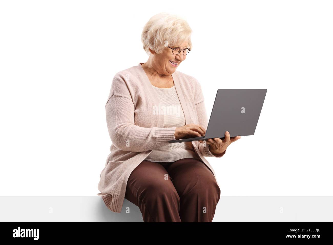 Donna anziana seduta su un pannello vuoto e che utilizza un computer portatile isolato su sfondo bianco Foto Stock