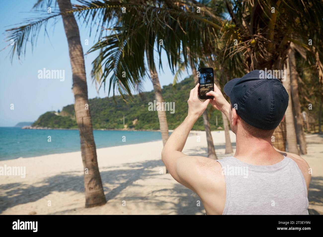Uomo che usa uno smartphone su una spiaggia di sabbia idilliaca. Vista posteriore del turista mentre fotografa palme in una destinazione tropicale. Foto Stock