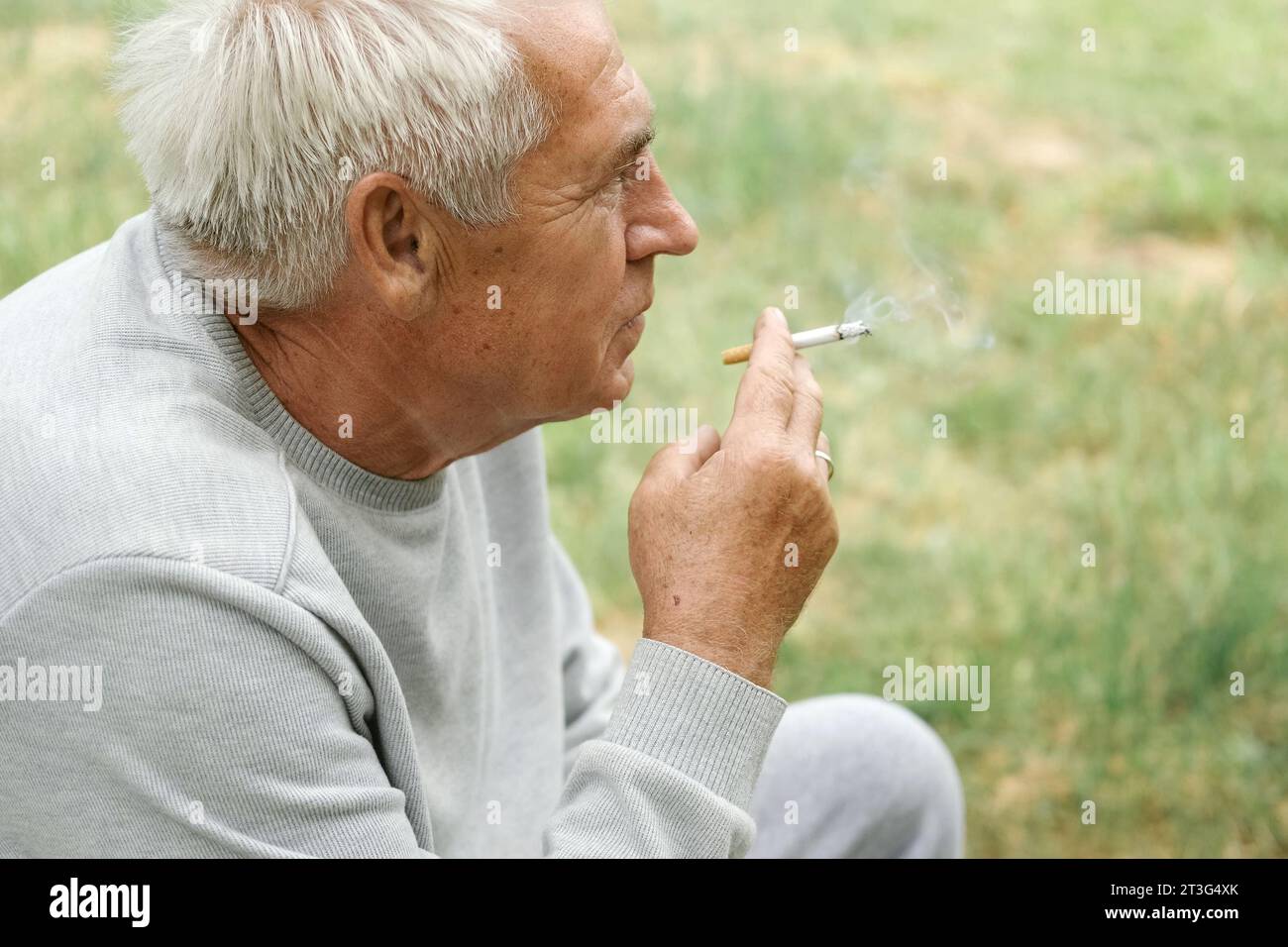Uomo anziano che accende una sigaretta all'aperto su sfondo verde. Vecchio uomo premuroso che fuma, guardando da parte. Faccia da vicino. Pensione rilassata Foto Stock