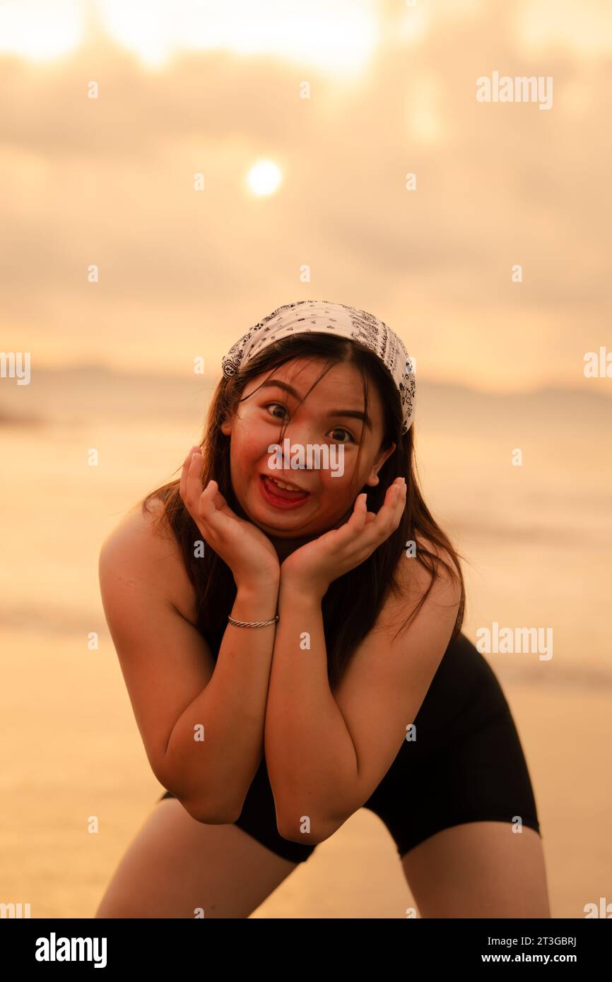 Un adolescente asiatico che indossa una bandanna bianca e una camicia nera con un'espressione goffa e felice sulla spiaggia al tramonto Foto Stock