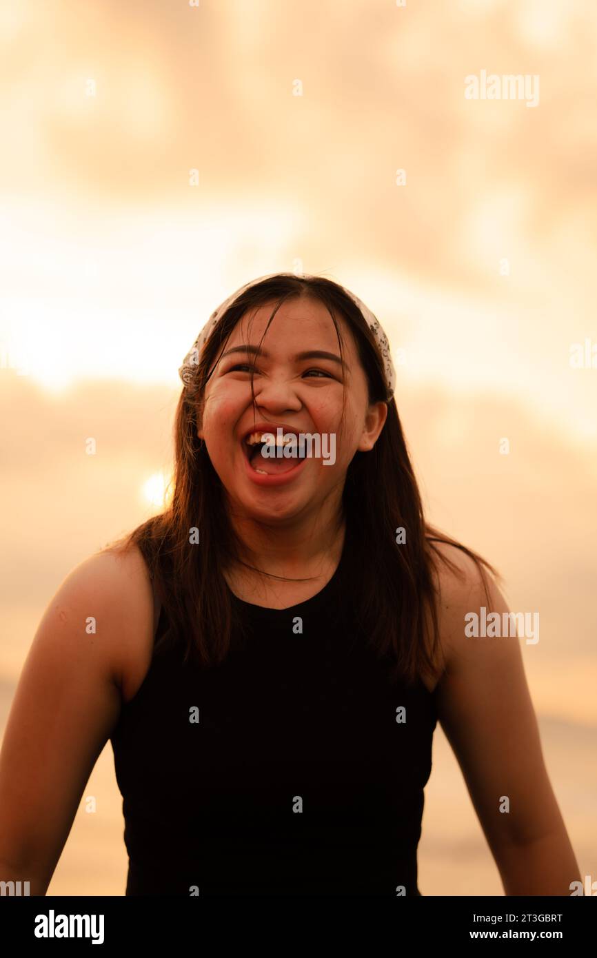 Un adolescente asiatico che indossa una bandanna bianca e una camicia nera con un'espressione goffa e felice sulla spiaggia al tramonto Foto Stock