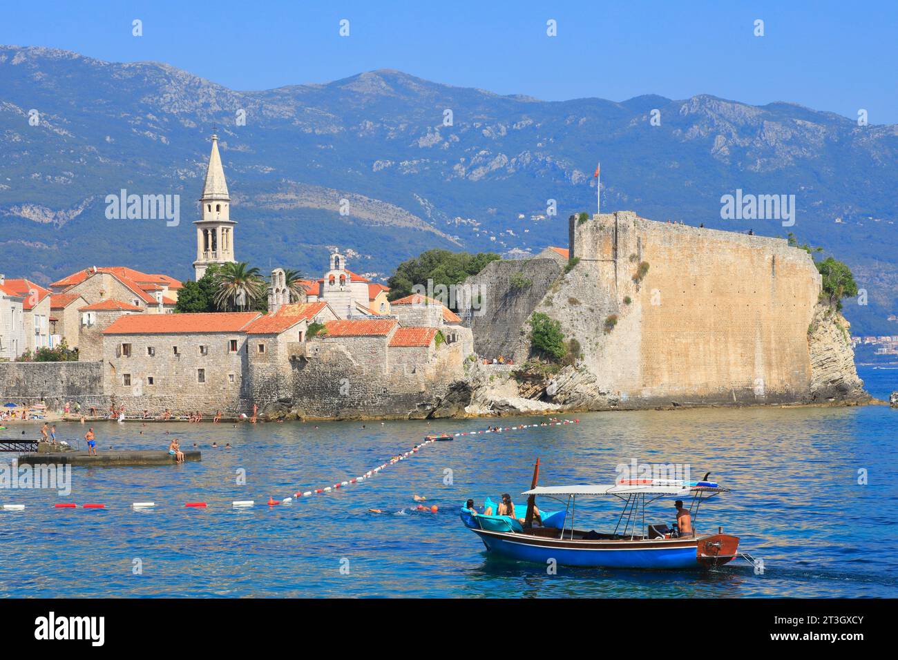 Montenegro, costa adriatica, Budva, città vecchia (Stari Grad) e St Chiesa di Giovanni (a sinistra) con la spiaggia e una barca turistica in primo piano Foto Stock