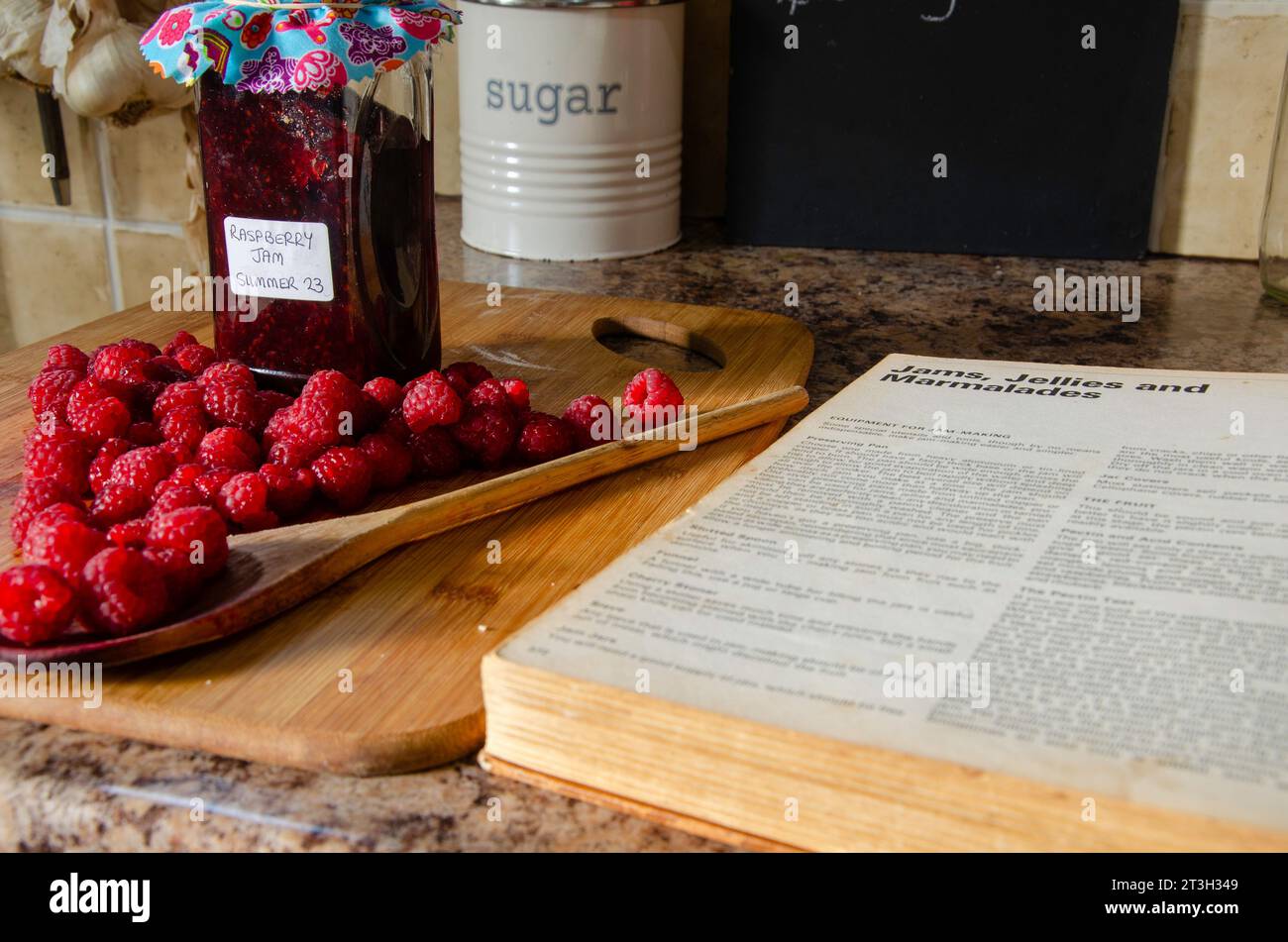 Lamponi, zucchero e un barattolo di marmellata insieme a un ricettario su un piano di lavoro Foto Stock