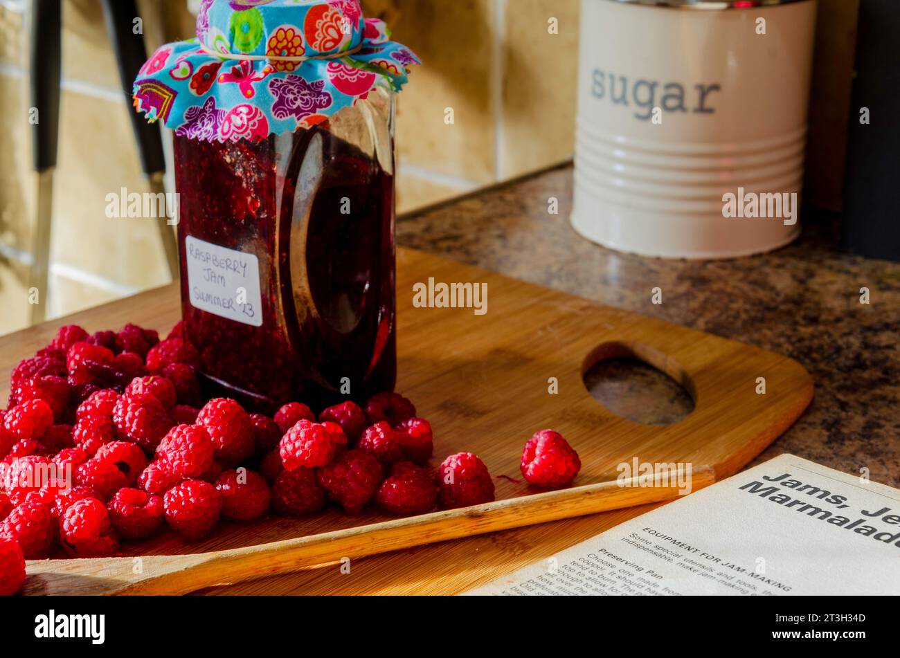 Lamponi, zucchero e un barattolo di marmellata insieme a un ricettario su un piano di lavoro Foto Stock