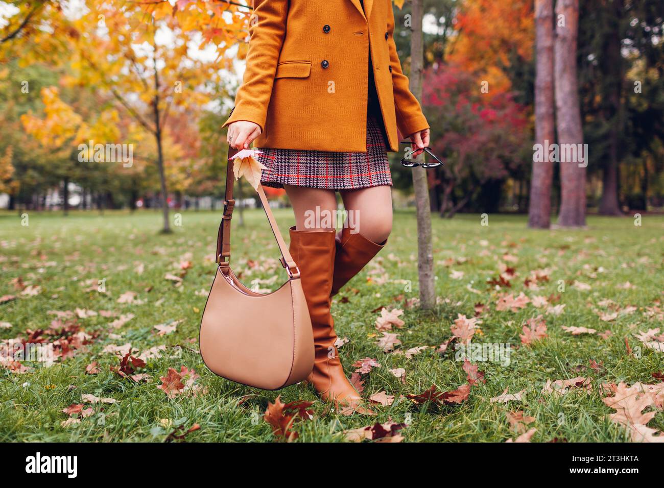 Stivali alti al ginocchio arancioni. Donna alla moda che indossa un blazer elegante minigonna a quadri che cammina nel parco autunnale con una borsa beige tra le foglie. Copia spazio Foto Stock