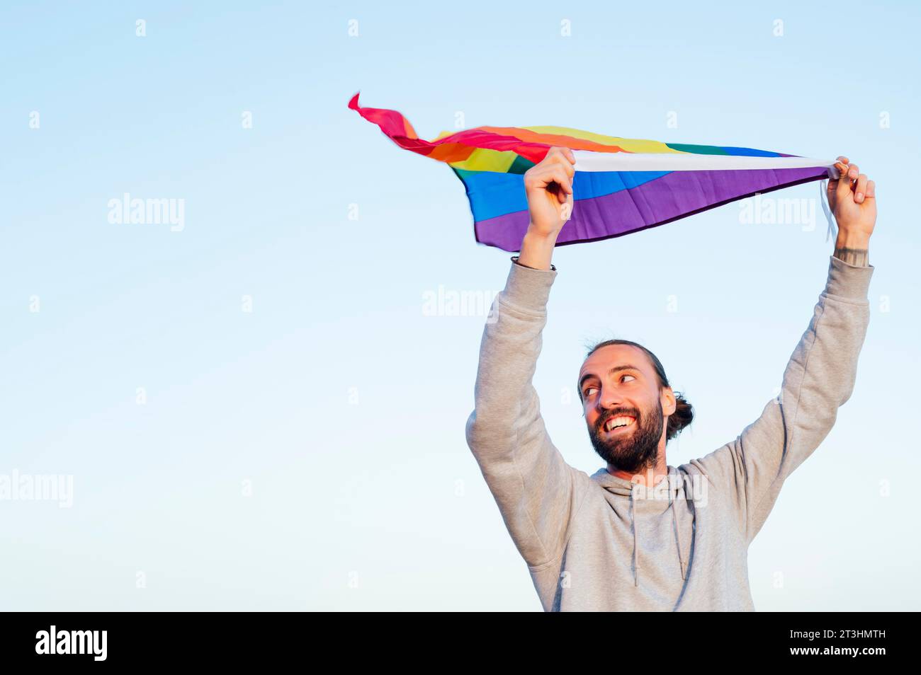 Ragazzo allegro con una bandiera arcobaleno lgbtq sulla spiaggia. Giovane che tiene una bandiera arcobaleno contro il cielo blu Foto Stock