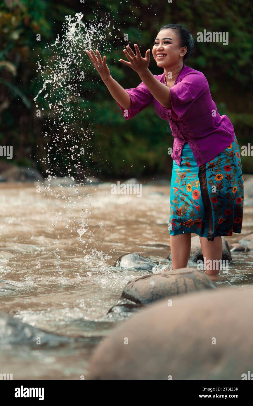 Una bella donna asiatica che si lava la mano e gioca con l'acqua mentre si trova vicino al fiume in un tradizionale vestito viola e gonna verde nel Foto Stock