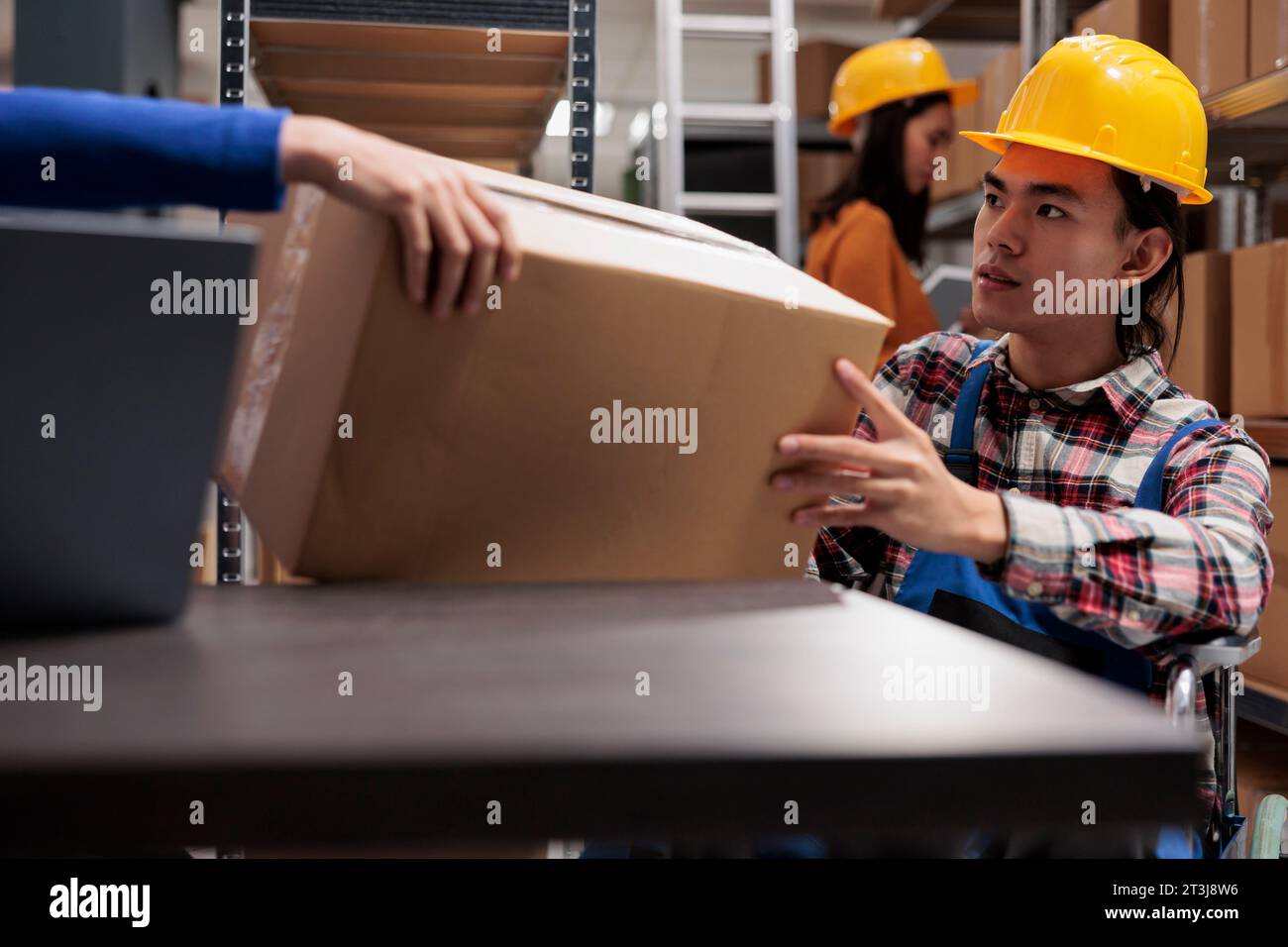 Commissionatore Storehouse su sedia a rotelle per l'evasione degli ordini e il prelievo di scatole di cartone dalle mani dei colleghi. Uomo asiatico con disabilità che gestisce le operazioni di servizio postale in un magazzino industriale Foto Stock