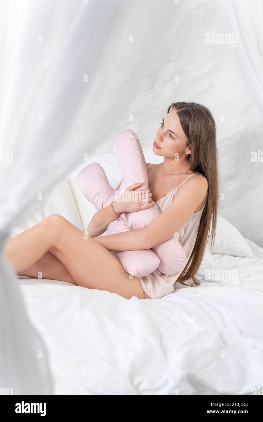 Bella ragazza con un cuscino di bellezza in mano siede a letto ed è triste. Foto verticale Foto Stock