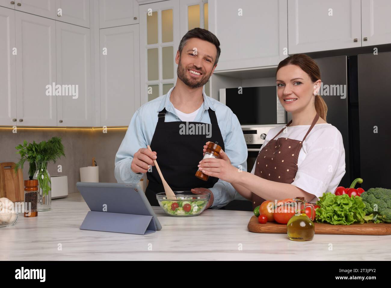 Coppia felice che legge la ricetta su un tablet mentre cucina in cucina. Libro culinario online Foto Stock
