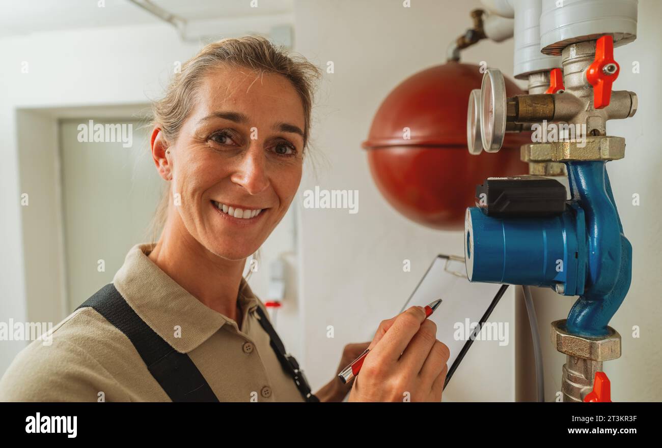 Donna sorridente ingegnere del riscaldamento in una sala caldaie a gas per il riscaldamento dell'impianto con lista di controllo su Appunti. Immagine del concetto di obbligo di sostituzione del riscaldatore a gas Foto Stock