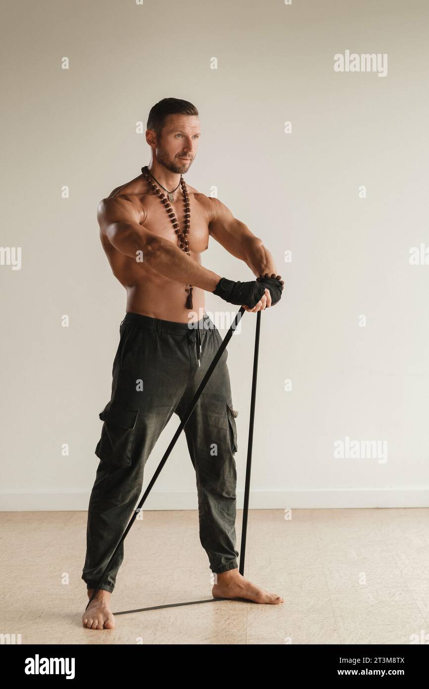 Un uomo con il torso nudo è impegnato nel fitness della forza usando un anello di gomma al chiuso. Foto Stock