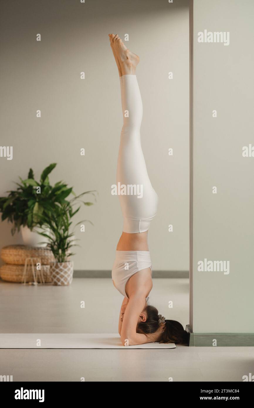 Una ragazza con vestiti bianchi fa yoga in piedi sulla testa al chiuso. Foto Stock