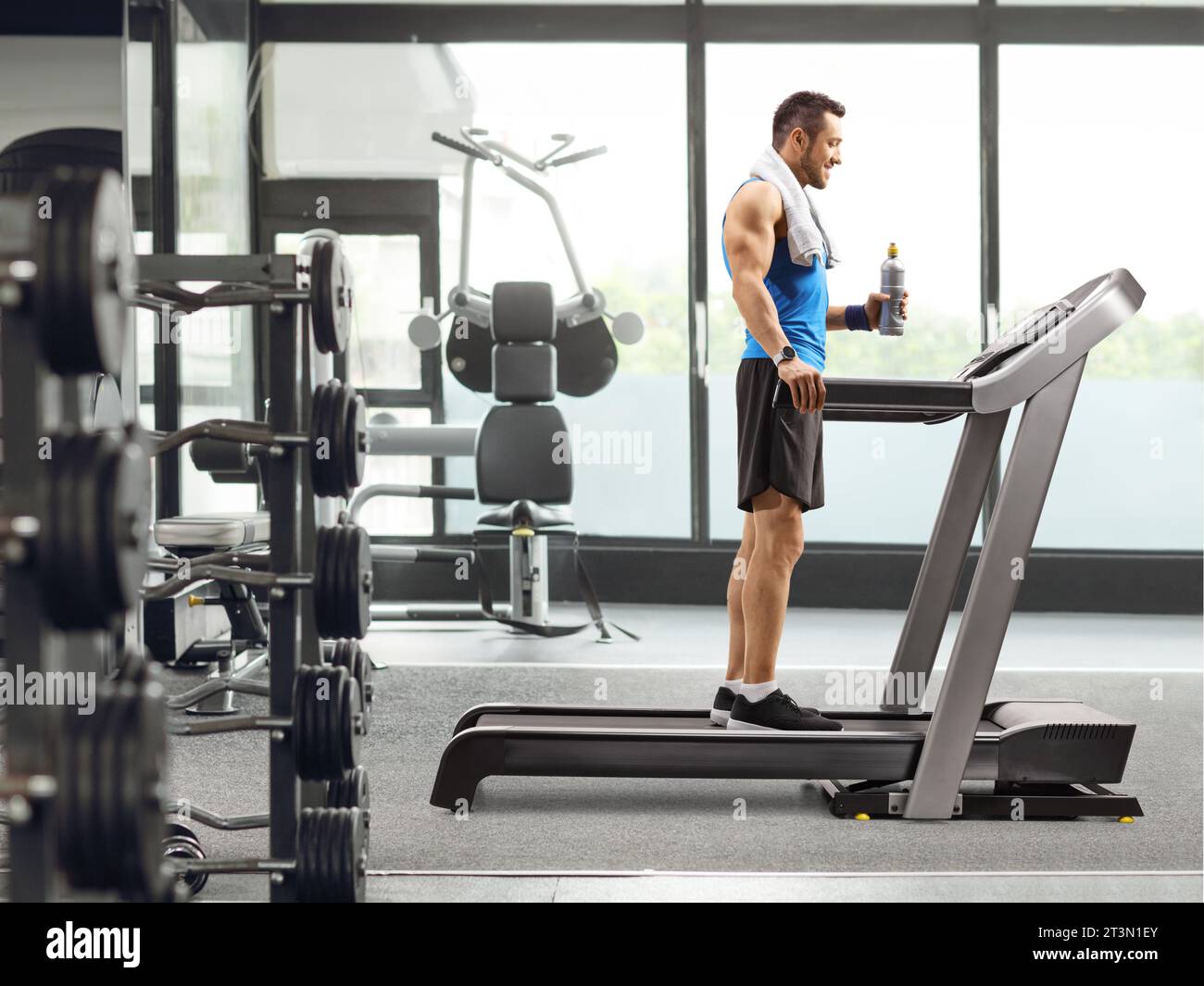 Foto a lunghezza intera di un uomo in forma in piedi su un tapis roulant con una bottiglia di bevanda in palestra Foto Stock