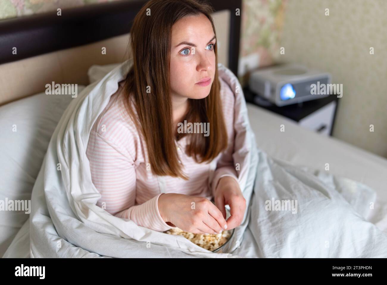 Una donna avvolta in una coperta che guarda un film sul suo letto usando un proiettore Foto Stock