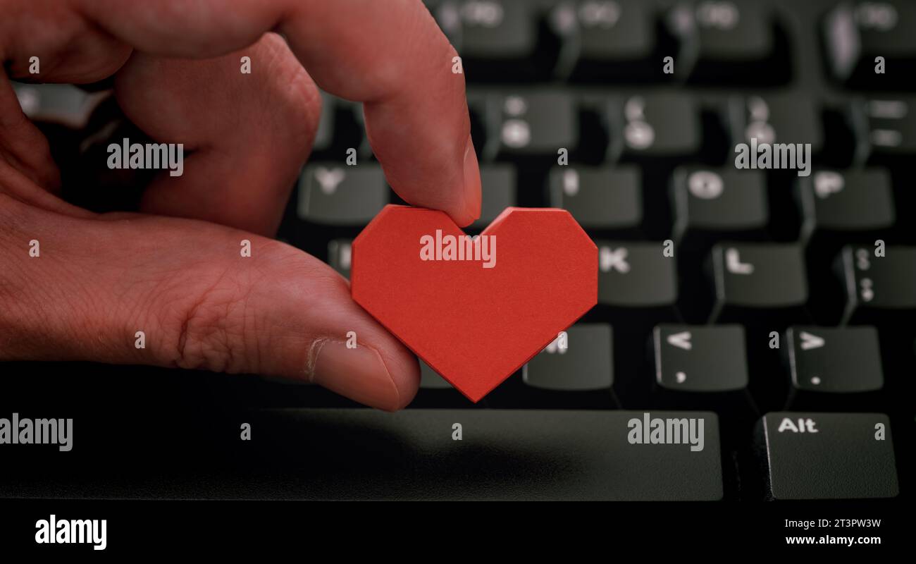 Un uomo che tiene in mano un cuore di carta origami rossa sulla tastiera nera del computer. Concetto di incontri online. Foto Stock