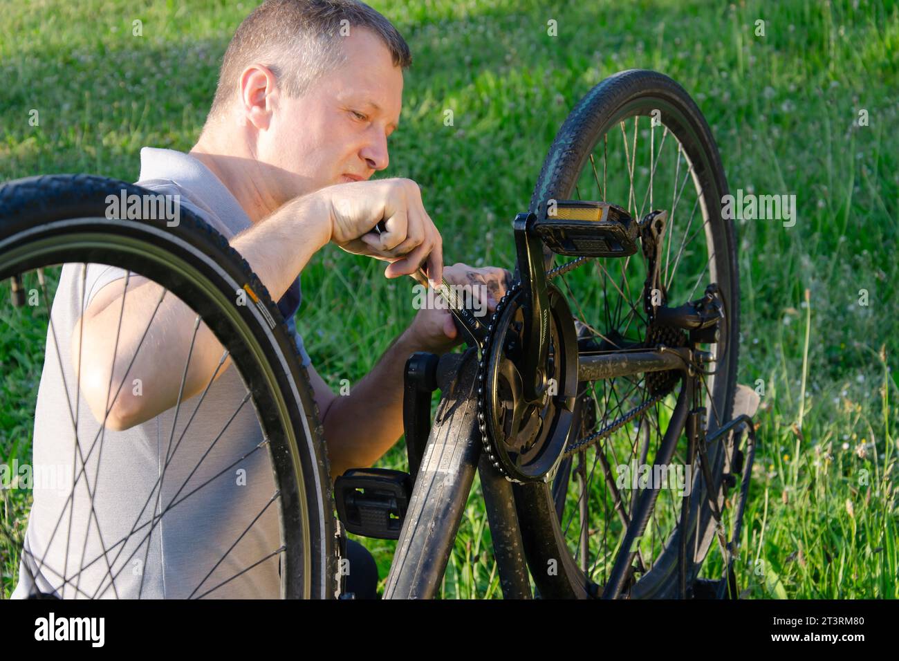 l'uomo sta riparando una catena di biciclette nel parco estivo. Indossa una t-shirt grigia. la bicicletta è posizionata diagonalmente nel telaio. lo spazio circostante è gre Foto Stock