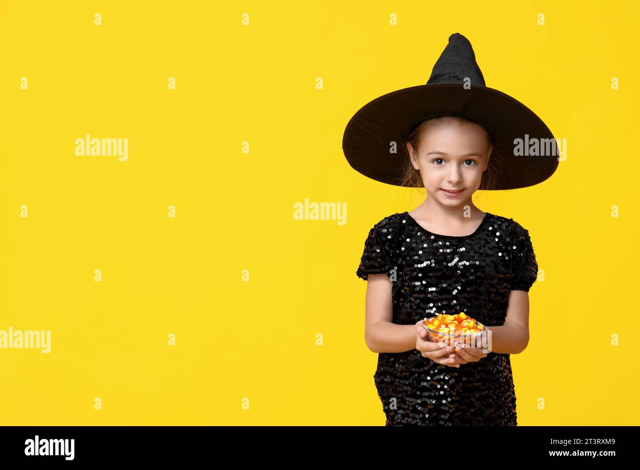 Graziosa bambina vestita per Halloween come una strega con una ciotola di gustose caramelle su sfondo giallo Foto Stock