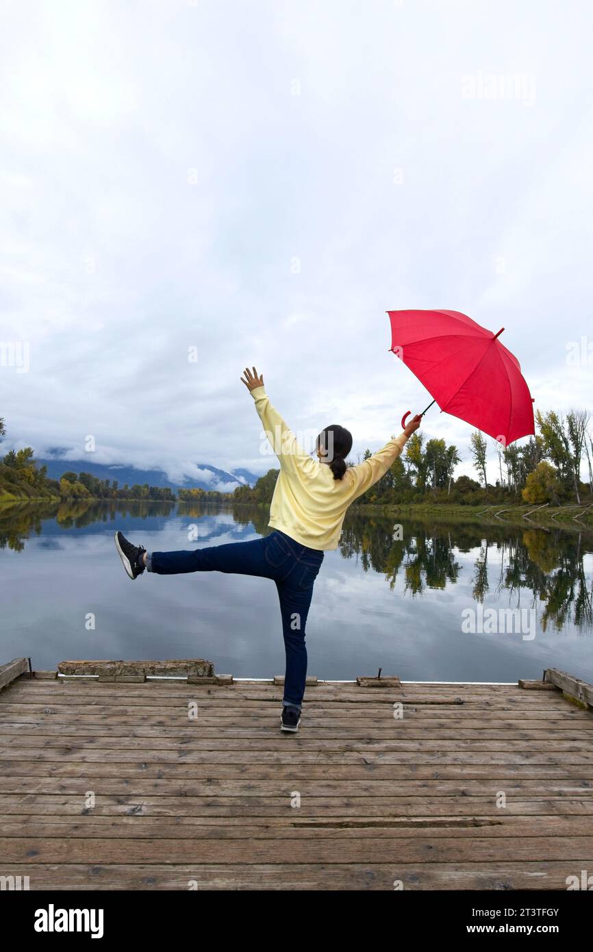 Una donna che tiene un ombrello rosso si mette in posa mentre si trova su un molo vicino al fiume Kootenai nell'Idaho settentrionale. Foto Stock