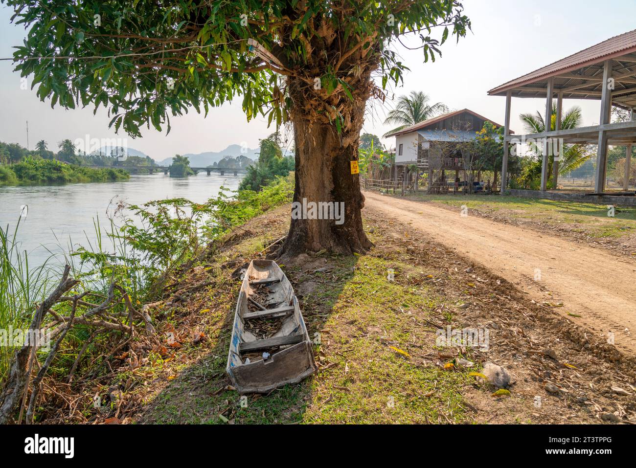 Tranquillo paesaggio fluviale rurale, antico ponte coloniale francese Don Det-Don Khon in lontananza, stretta strada sterrata che passa davanti a un albero alberato a strapiombo Foto Stock