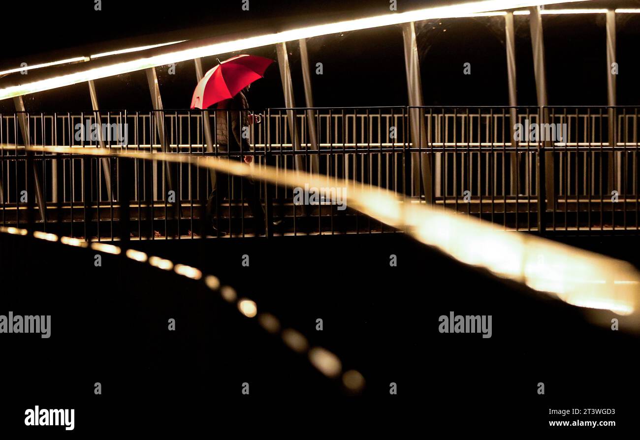 Riedlingen, Germania. 27 ottobre 2023. Un uomo con un ombrello cammina al mattino sotto la pioggia su un ponte illuminato sul Danubio. Crediti: Thomas Warnack/dpa/Alamy Live News Foto Stock