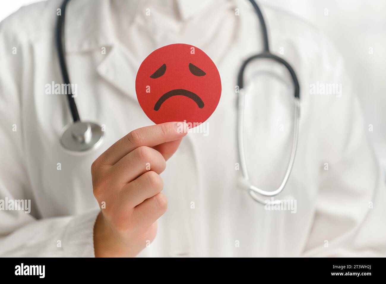 Primo piano di una giovane medico in uniforme bianca con stetoscopio che regge un'emoticon rossa del viso arrabbiato. Intelligenza emotiva, equilibrio controllo emozionale, io Foto Stock