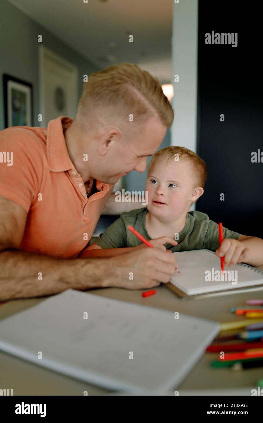 Padre che assiste il figlio con la sindrome di Down nello studio al tavolo da pranzo Foto Stock