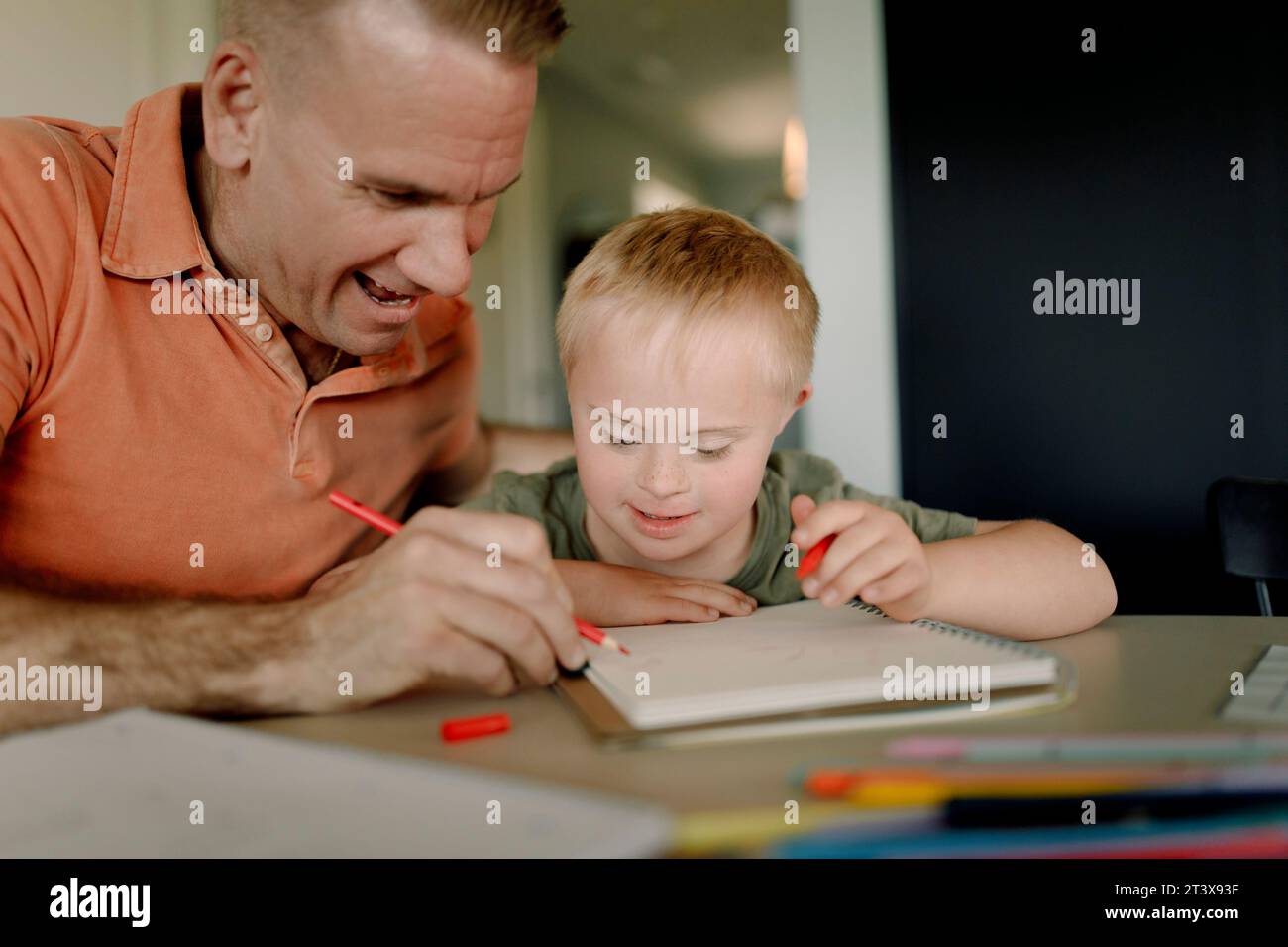 Padre che assiste il figlio con la sindrome di Down nel scrivere un libro a tavola Foto Stock