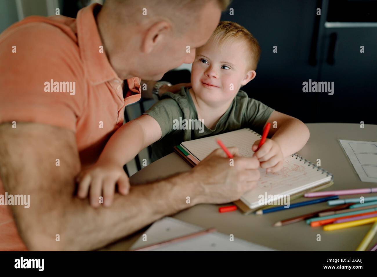 Ragazzo con sindrome di Down che guarda il padre mentre disegna sul libro Foto Stock