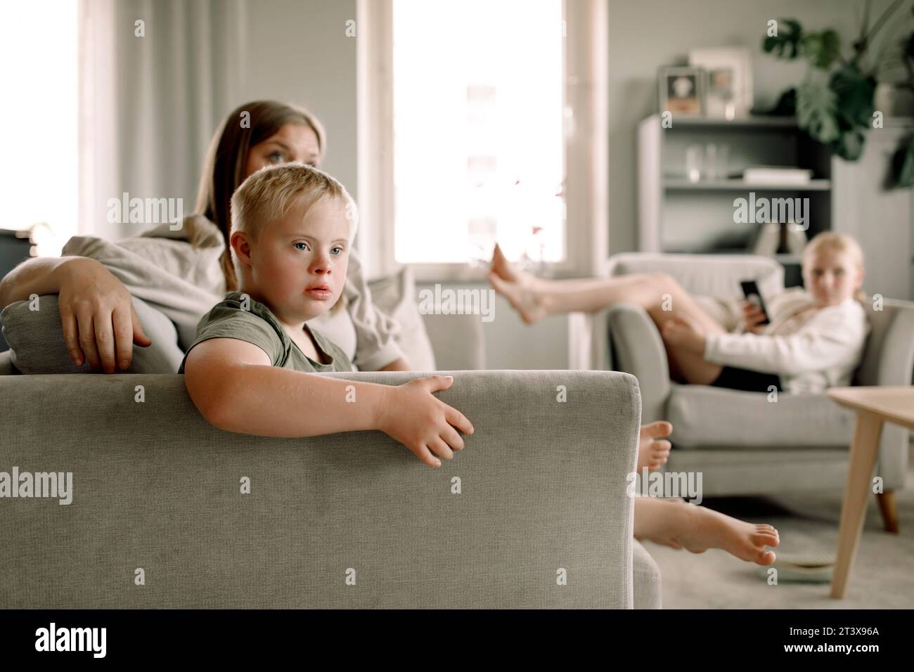 Ragazzo con sindrome di Down che guarda la TV dalla madre seduta sul divano a casa Foto Stock