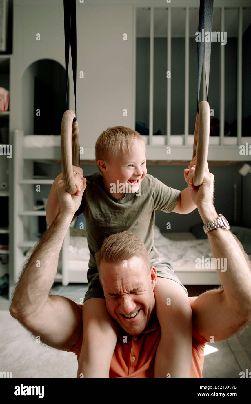 Uomo che trasporta un figlio con sindrome di Down sulle spalle mentre tiene anelli ginnici in camera da letto Foto Stock