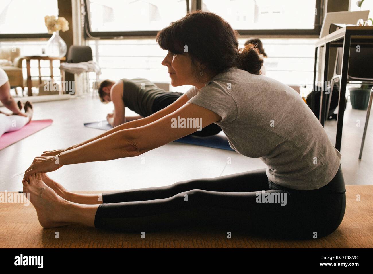 Vista laterale completa di una donna d'affari matura e flessibile che tocca le dita dei piedi mentre si pratica yoga sul tappetino sul posto di lavoro Foto Stock