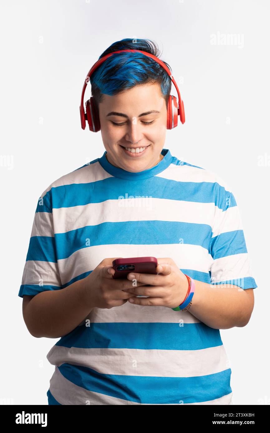 Una persona non binaria con capelli blu brillante, che indossa cuffie, ascolta la musica sul proprio smartphone, mostrando un'espressione pacifica e contenta. GA Foto Stock