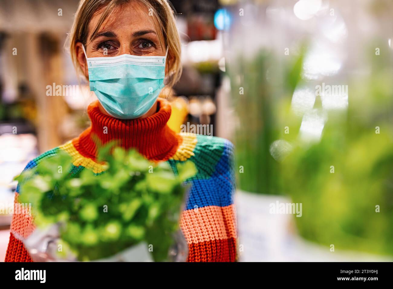 la donna che indossa una maschera facciale azzurra esamina il basilico fresco in un vivace negozio di alimentari o supermercato, circondato da una vasta gamma di prodotti. Sicurezza corona Foto Stock
