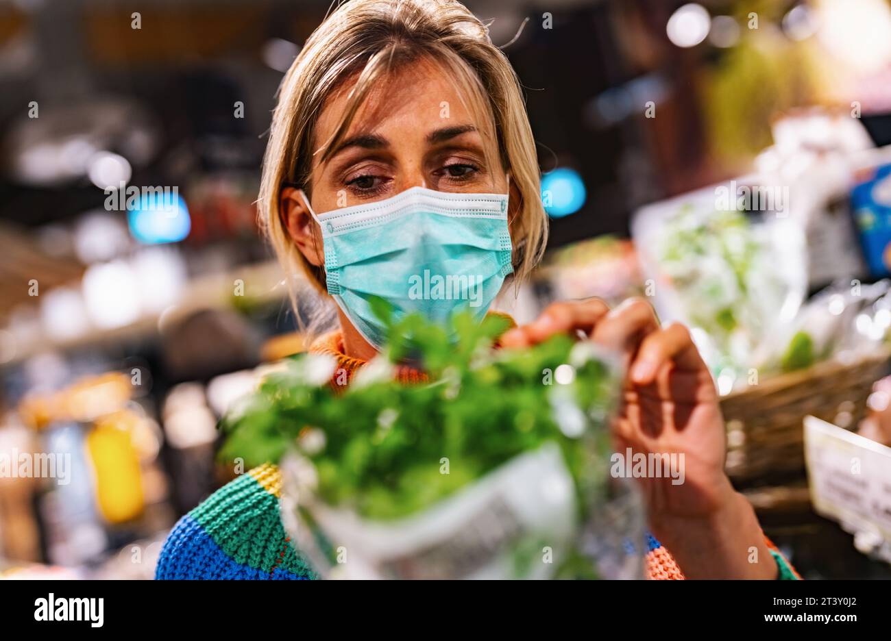 la donna che indossa una maschera facciale azzurra esamina il basilico fresco in un vivace negozio di alimentari o supermercato, circondato da una vasta gamma di prodotti. Sicurezza corona Foto Stock