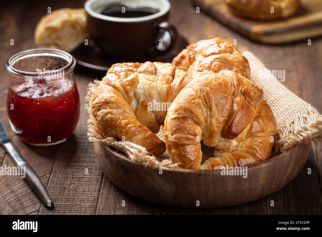 Croissant freschi in una ciotola di legno con tazza di caffè e barattolo di confetture di fragole sullo sfondo Foto Stock