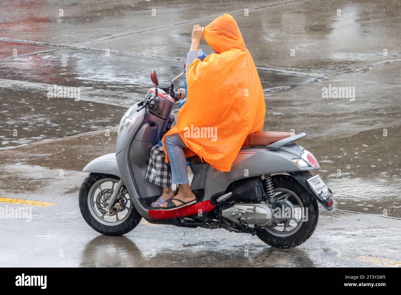 Una donna con un bambino cavalca una moto per strada sotto la pioggia battente, Thailandia Foto Stock