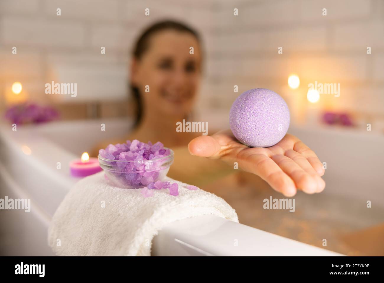 donna che si rilassa nella vasca da bagno a lume di candela e utilizza una bomba da bagno con cristalli di sale marino per idratare la pelle del corpo. centro benessere domestico, cura della pelle Foto Stock