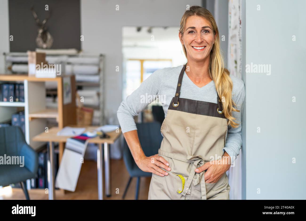 allegra donna bionda con top grigio e grembiule beige in un'area di lavoro sartoriale. Ha un metro a nastro e una forbice nella tasca del grembiule ed è S Foto Stock
