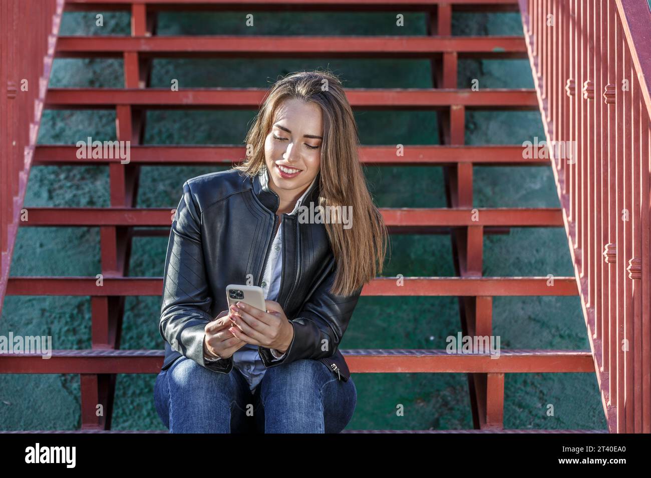 Giovane donna attraente con i capelli lunghi e chiari che indossa una giacca in pelle seduta su una scala rossa e chiacchierata sui social media tramite un moderno telefono cellulare Foto Stock