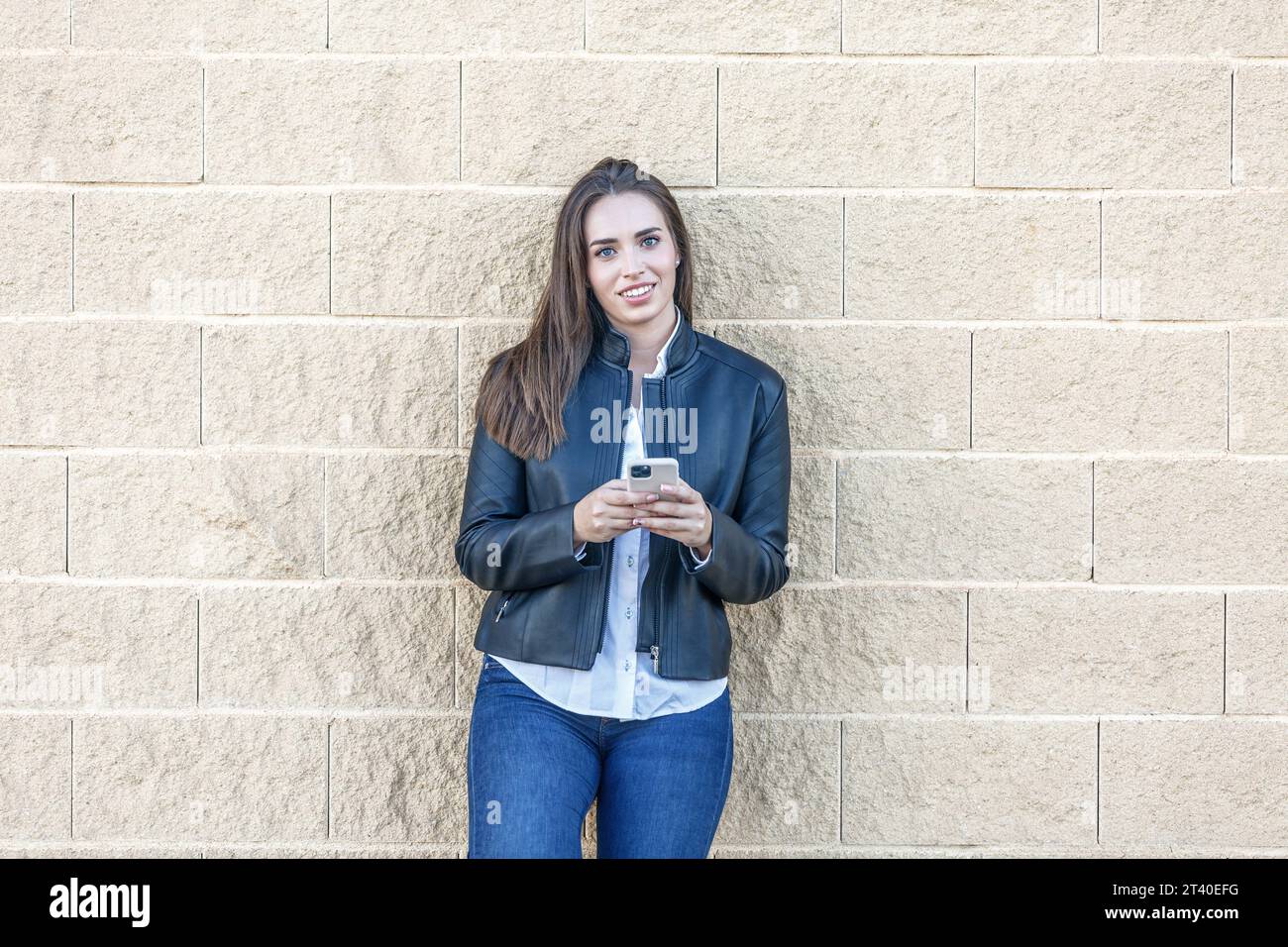 Giovane donna di 20 anni sorridente guardando al futuro, vestita da una giacca di pelle che regge uno smartphone appoggiato a una parete di mattoni Foto Stock