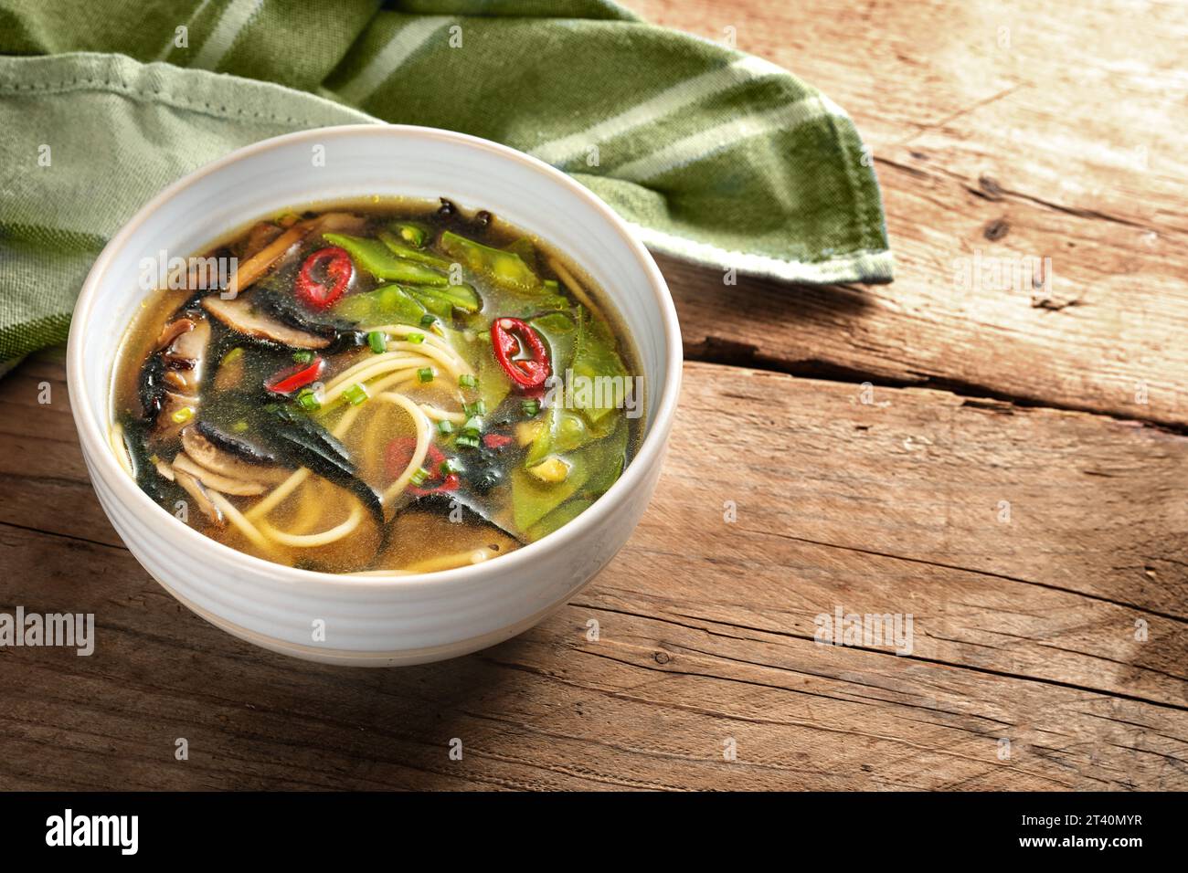 Zuppa di verdure con spaghetti, miso, alghe nori, piselli da neve, zenzero e peperoncino in una ciotola e un asciugamano verde su un tavolo rustico di legno, spazio per copiatrici, Foto Stock