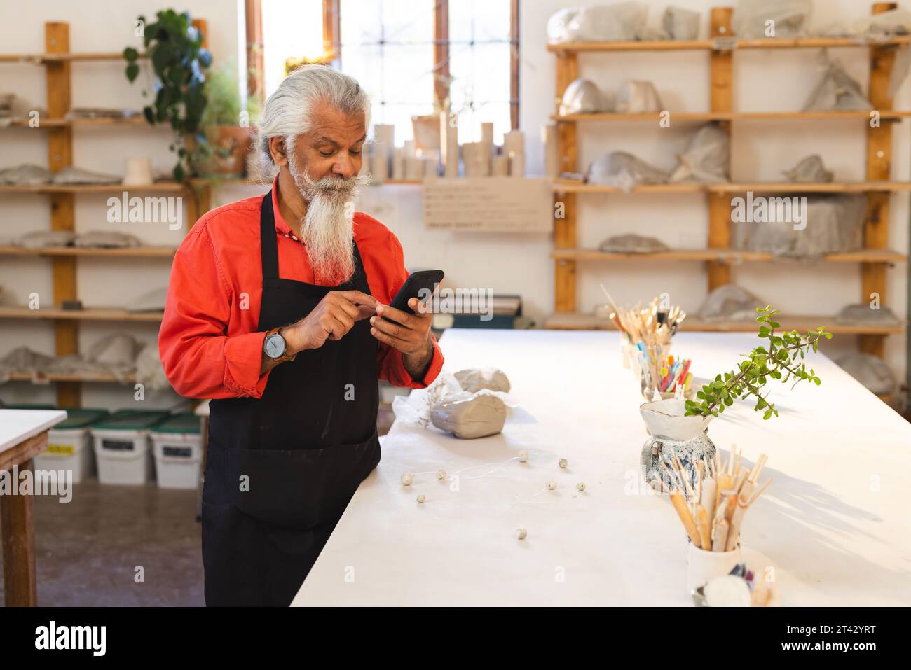 vasaio esperto birazziale con barba lunga con lo smartphone in uno studio di ceramica Foto Stock
