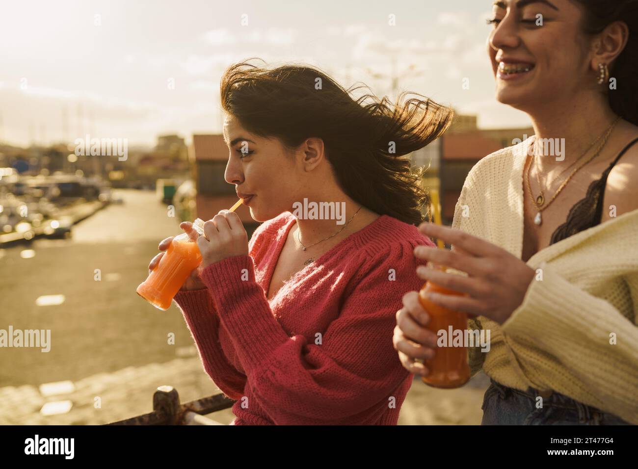 Due amici condividono un momento delizioso al porto, con il vento che si stringe i capelli. I sorrisi genuini e le bevande vivaci catturano l'essenza di un perfec Foto Stock