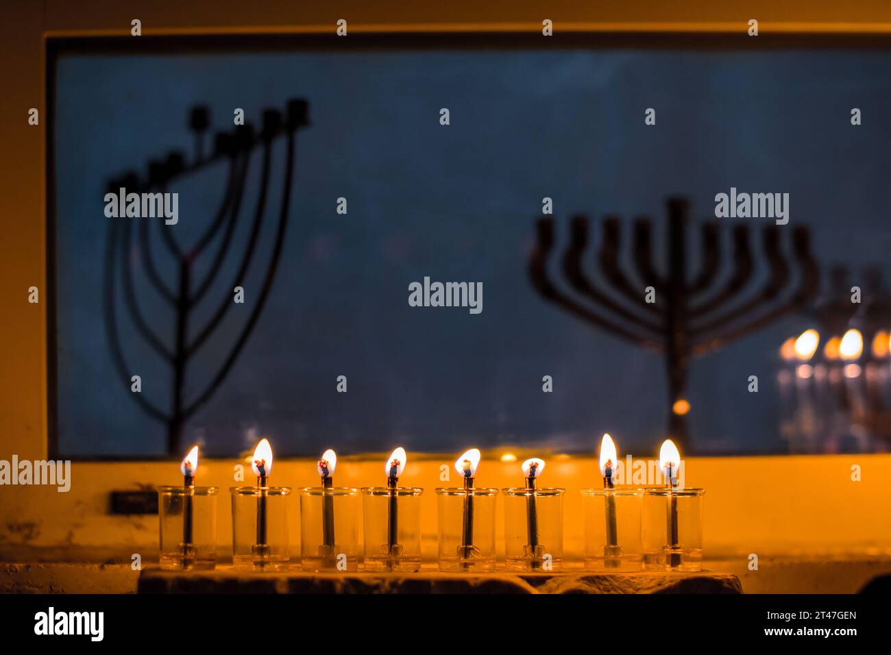 Le fiale di vetro contengono olio e stoppini brucianti in una menorah luminosa sotto una finestra con menorah non illuminate nell'ottava notte del festival ebraico Foto Stock