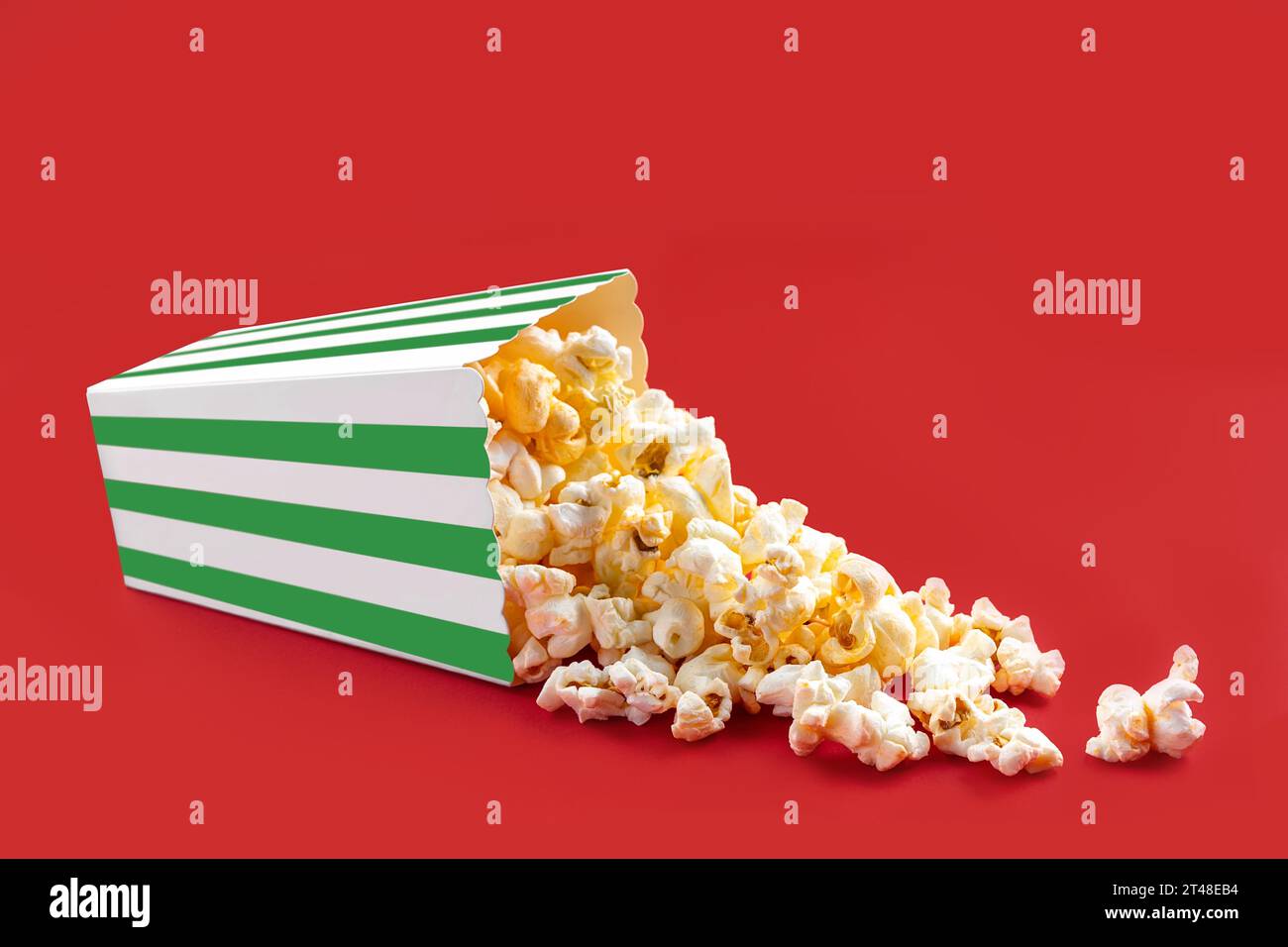 Gustosi popcorn al formaggio che cadono da una scatola di cartone a righe verdi o da un secchio, isolato su sfondo rosso. Dispersione di semi di popcorn. Fast food, snack. Foto Stock